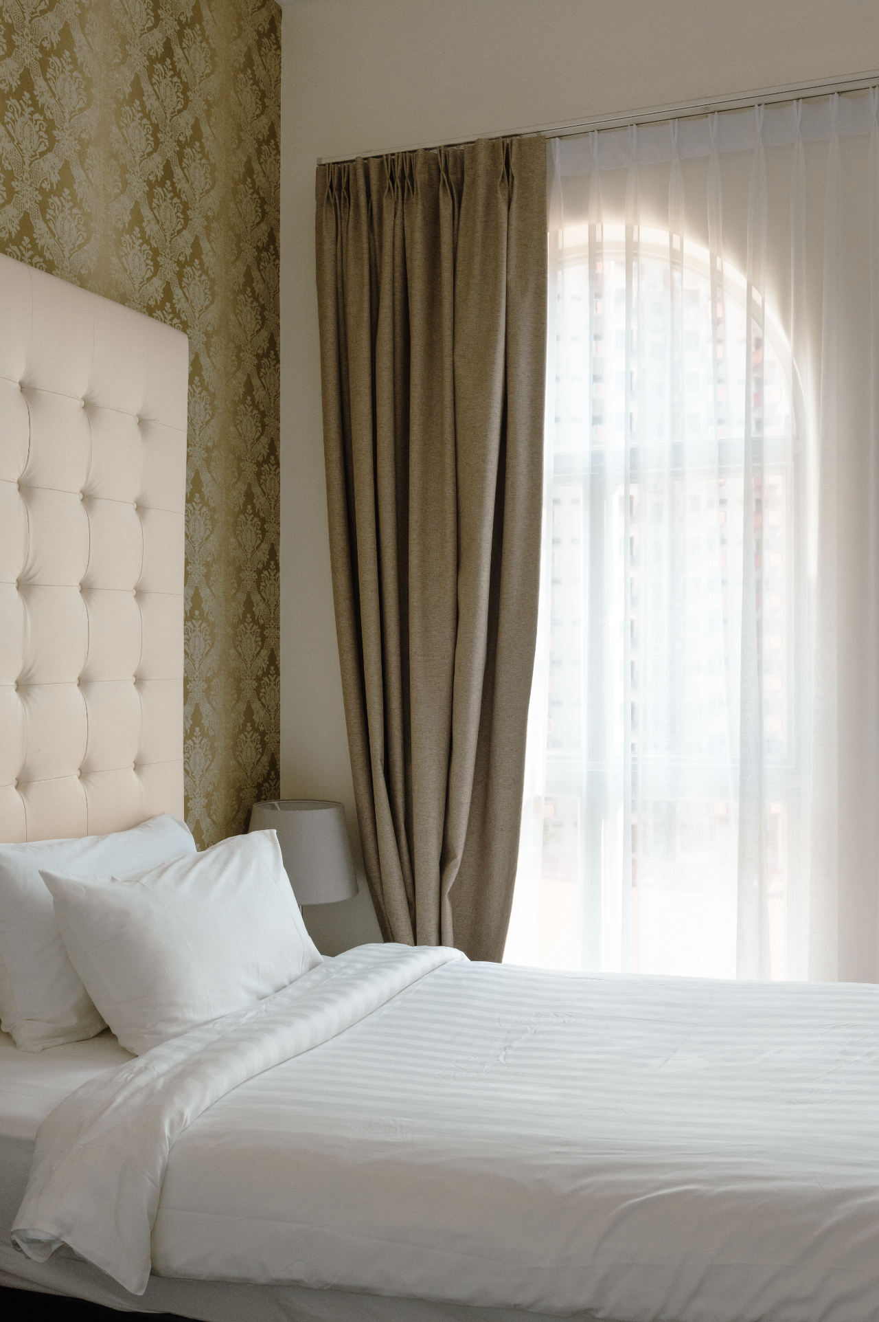 A neatly made bed with white bedding and pillows, a beige headboard, and a small white bedside lamp. Behind the bed is a wall with gold patterned wallpaper and a large window with sheer white curtains and beige drapes, showing a view of tall buildings outside.
