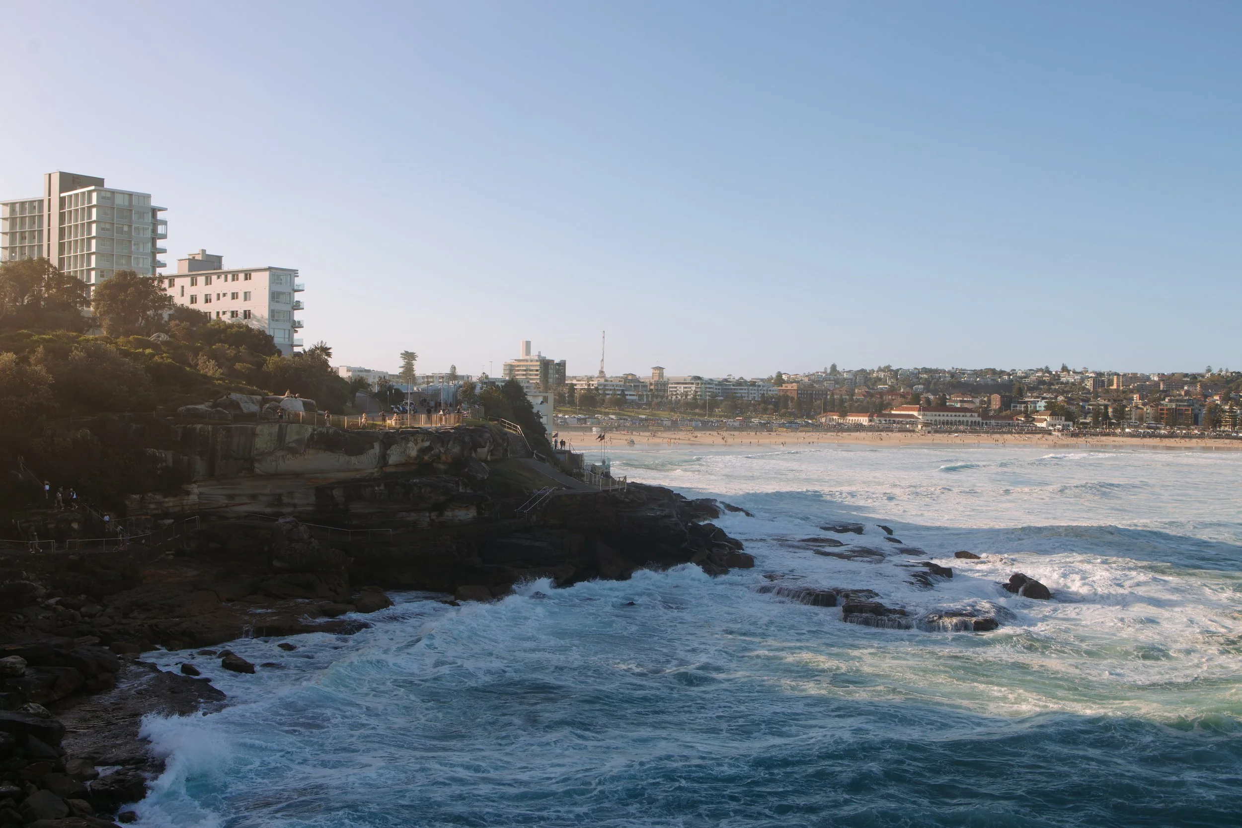 View of a beach with ocean waves crashing against rocks in the foreground and a city skyline with tall buildings and houses in the background under a clear blue sky.