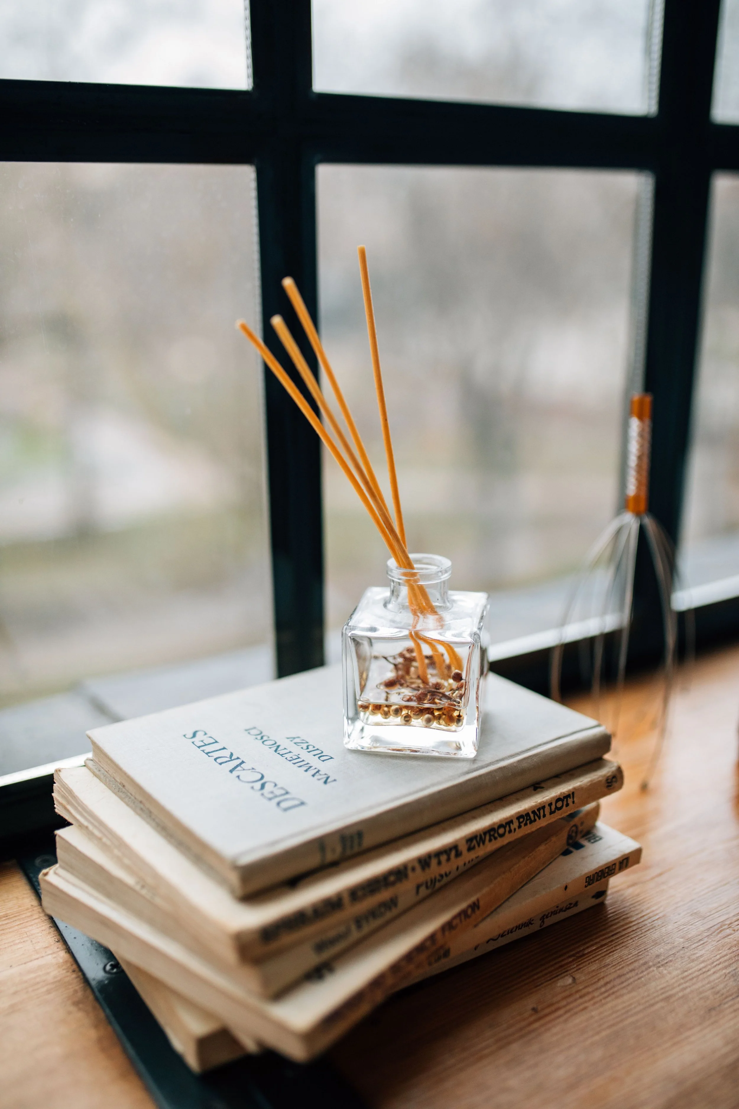 Stack of books with a glass jar containing incense sticks and a wooden whisk, placed on a wooden table near a window.