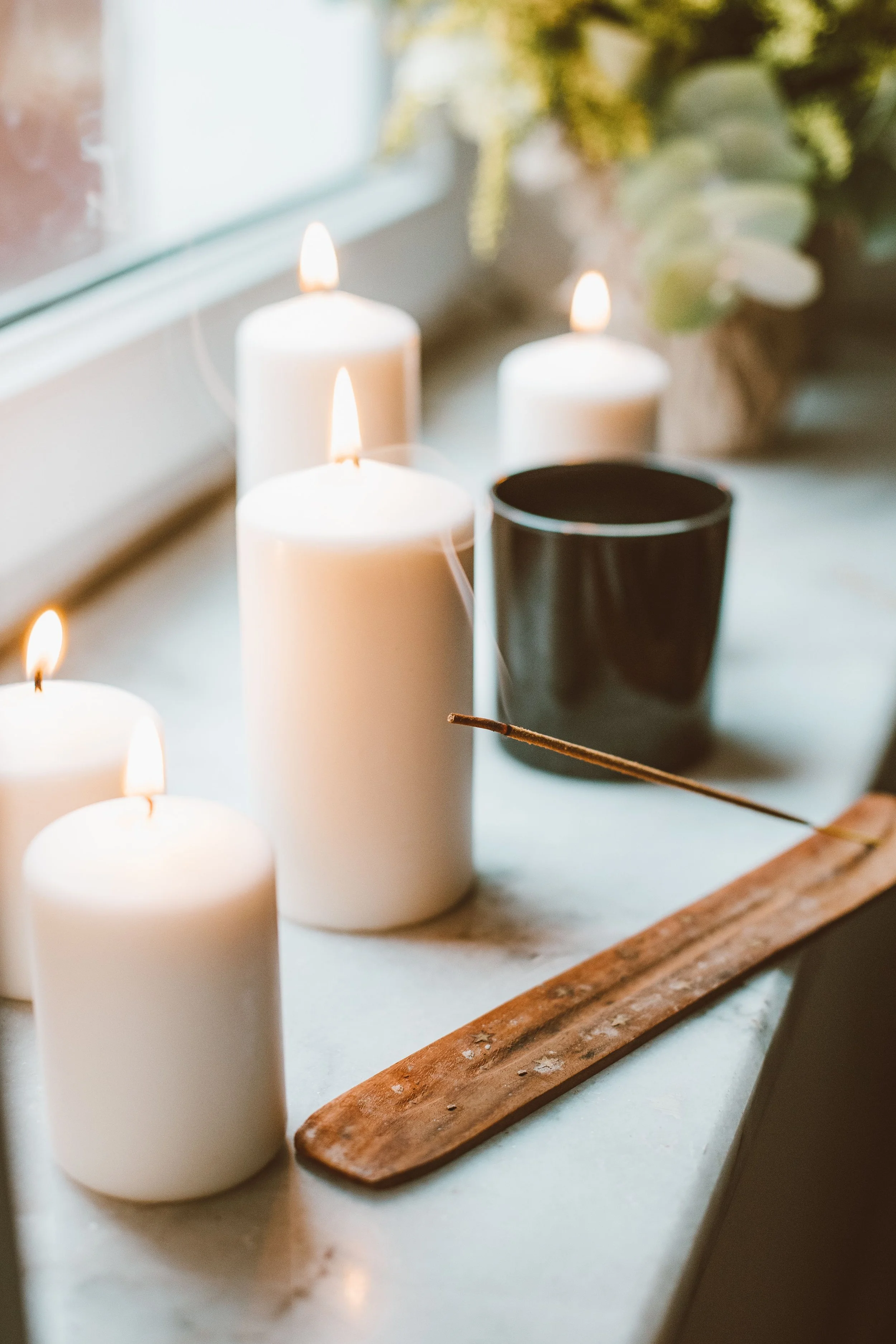 Four white candles, one in a black holder, are on a windowsill, with a wooden tray and some flowers in the background.