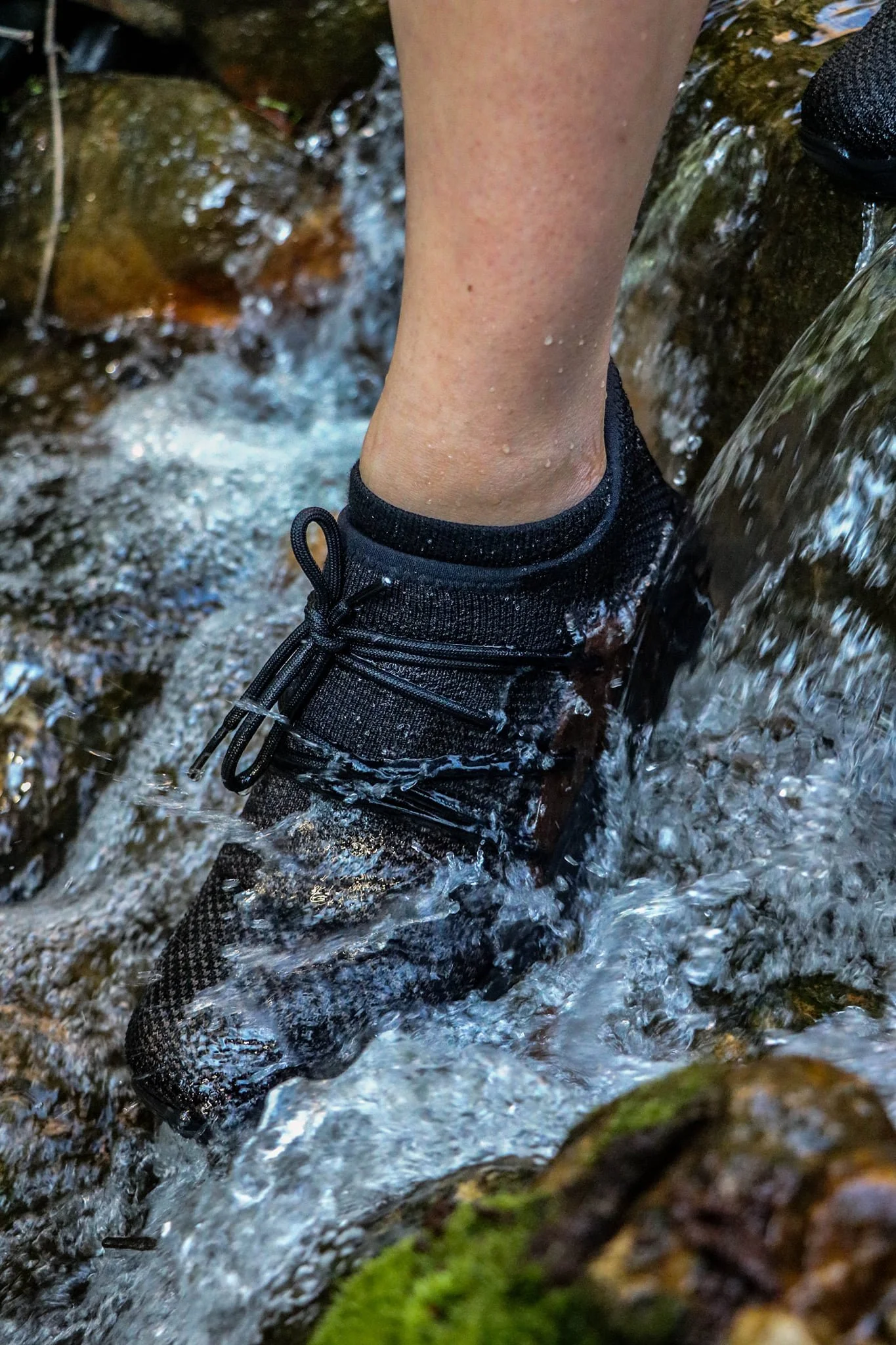 A person's leg and foot wearing a black hiking shoe stepping through a stream of water with rocks and moss around.