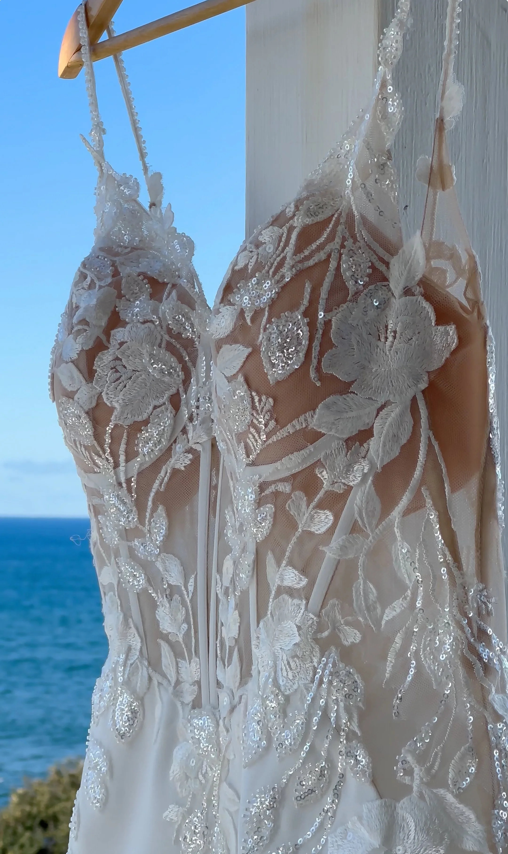Close-up of a wedding dress with intricate white floral embroidery and sequins, hanging outdoors against a backdrop of blue sky and ocean.
