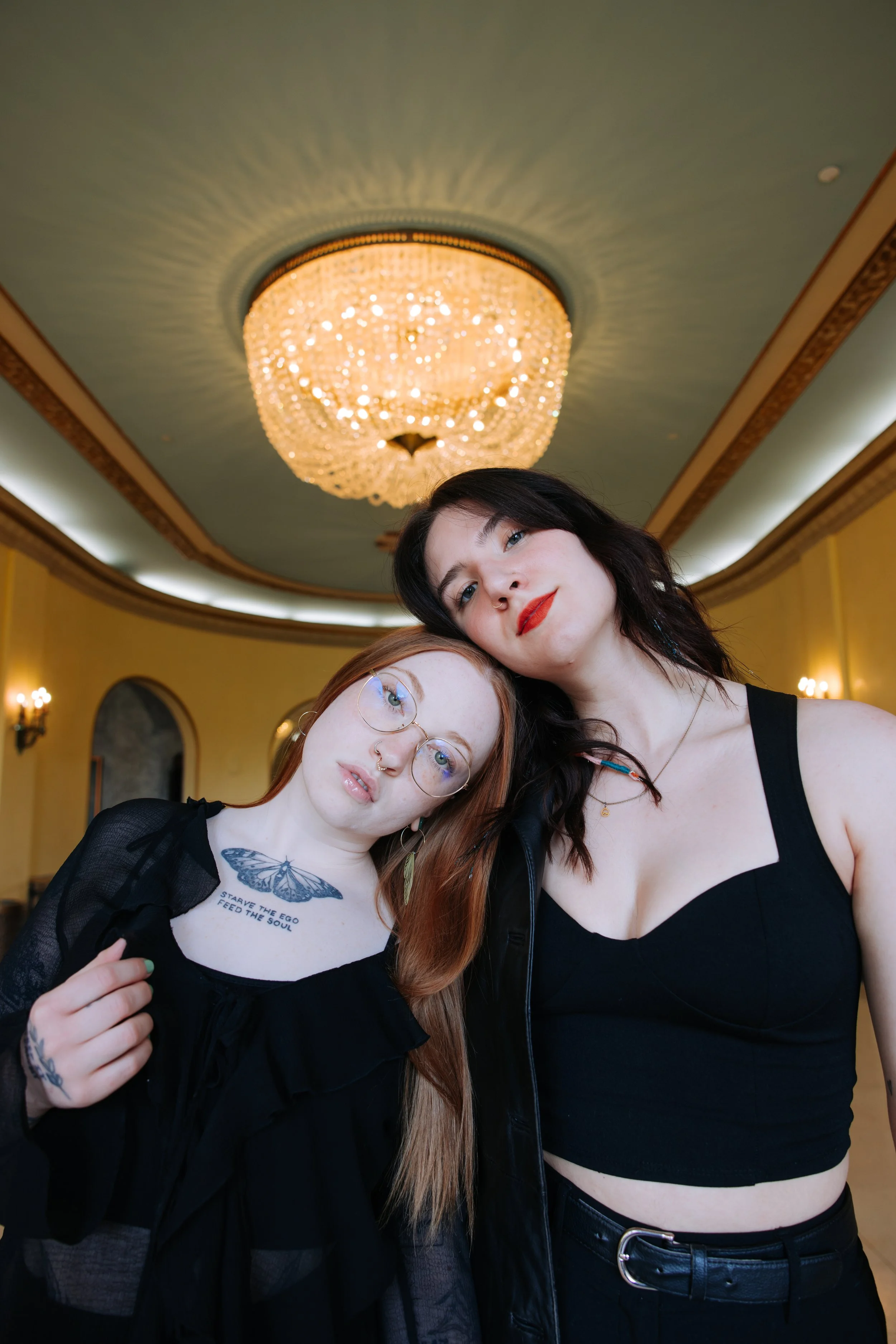 Two women posing in front of a chandelier in an ornate room. One has red hair, glasses, and tattoos, and the other has dark hair and red lipstick.