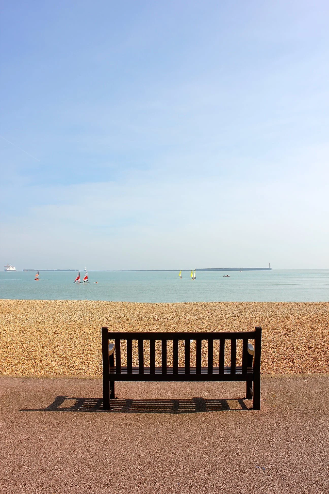 Empty park bench facing a sandy beach and calm sea with sailboats and a ship, under a partly cloudy sky.