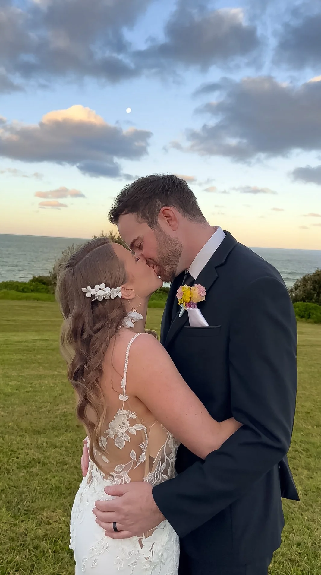 A bride and groom sharing a kiss outdoors by the ocean at sunset, with cloudy sky and grassy landscape in the background.