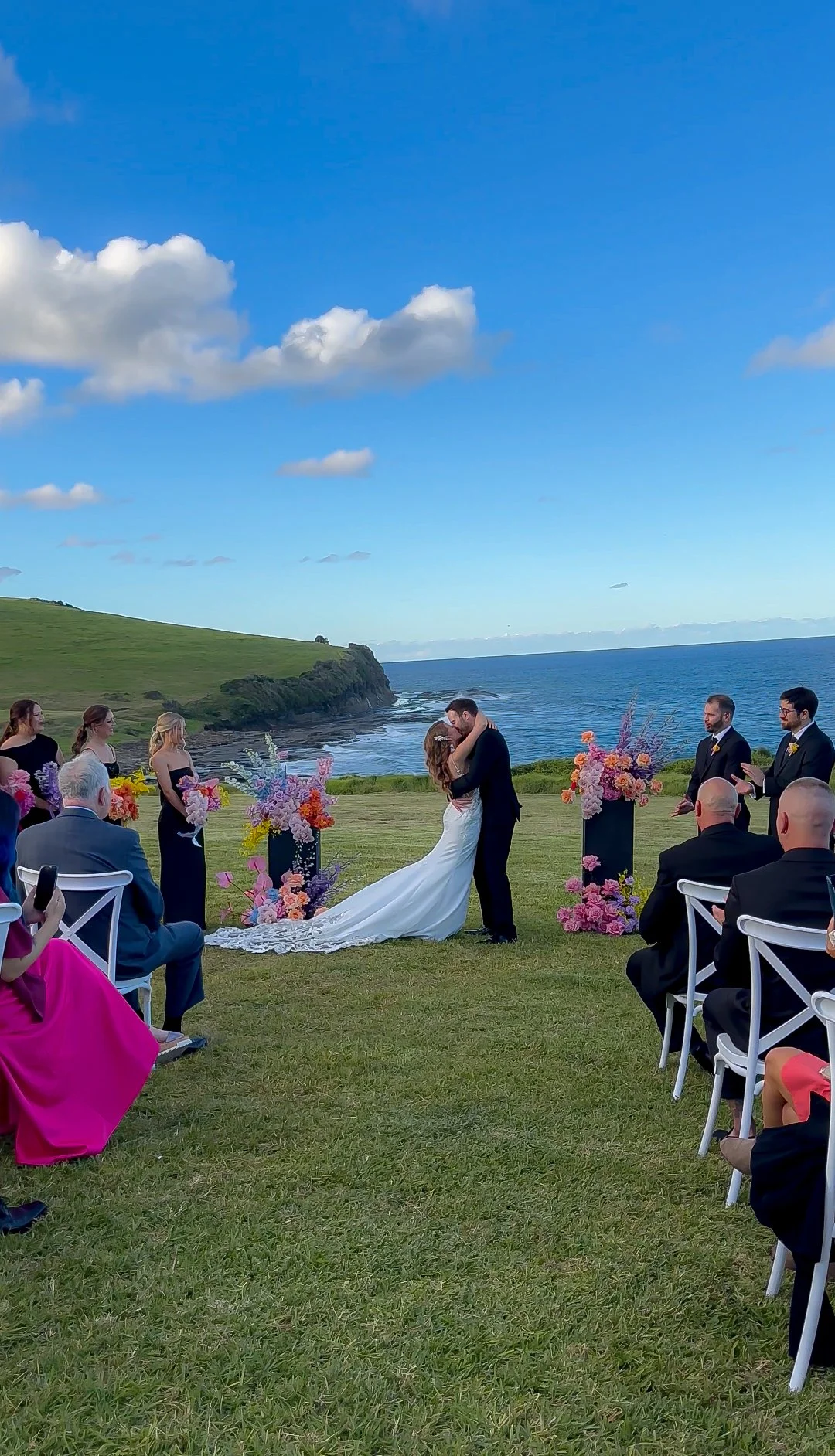 A bride and groom kiss during an outdoor wedding ceremony on a grassy area near the ocean, with a blue sky and clouds overhead. Guests sit on white chairs, and floral arrangements decorate the aisle.