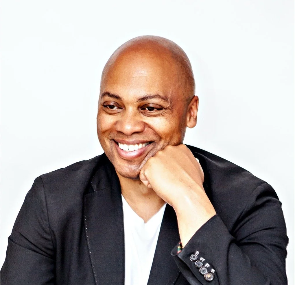 Close-up portrait of a smiling bald man in a black blazer, resting his chin on his hand against a plain white background.