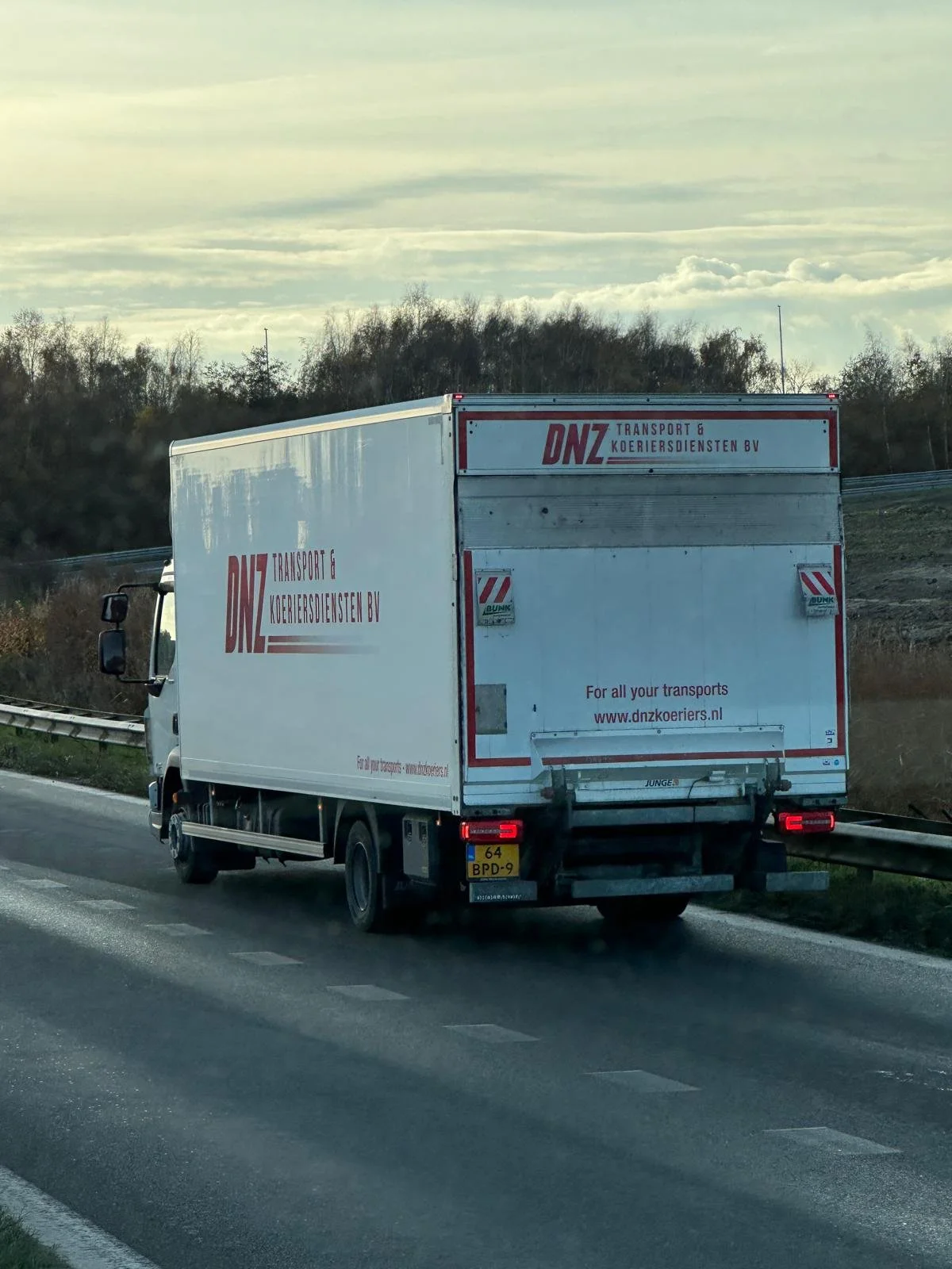 Een witte vrachtwagen met de logo's en naam van DNZ Transport en Koeriersdiensten BV rijdt op een autosnelweg met een bos en wolken in de achtergrond.