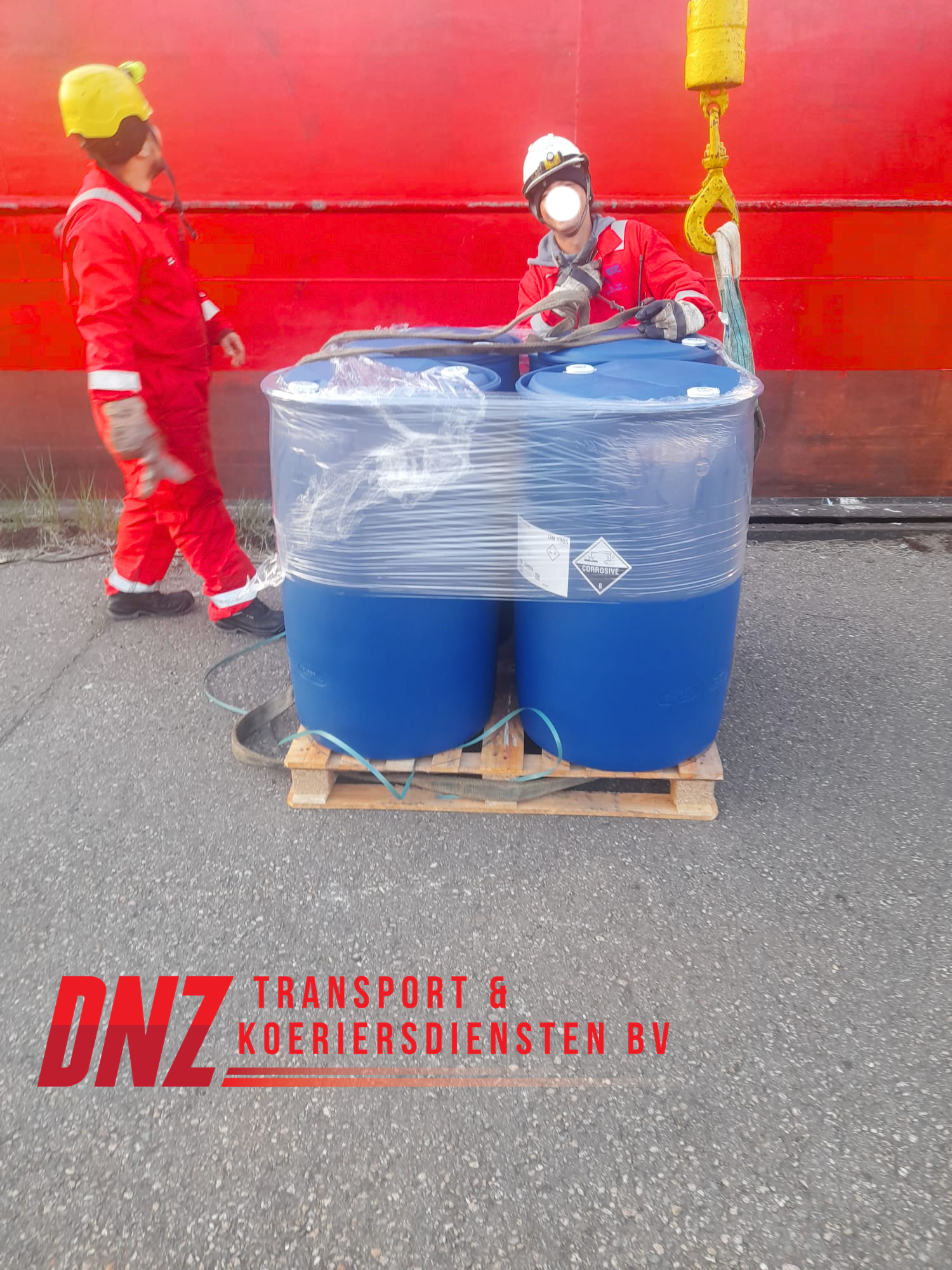 Twee mensen in veiligheidskleding en helmen staan naast grote blauwe vaten die op een houten pallet staan. Het lijkt een industriële setting met een rode muur op de achtergrond.