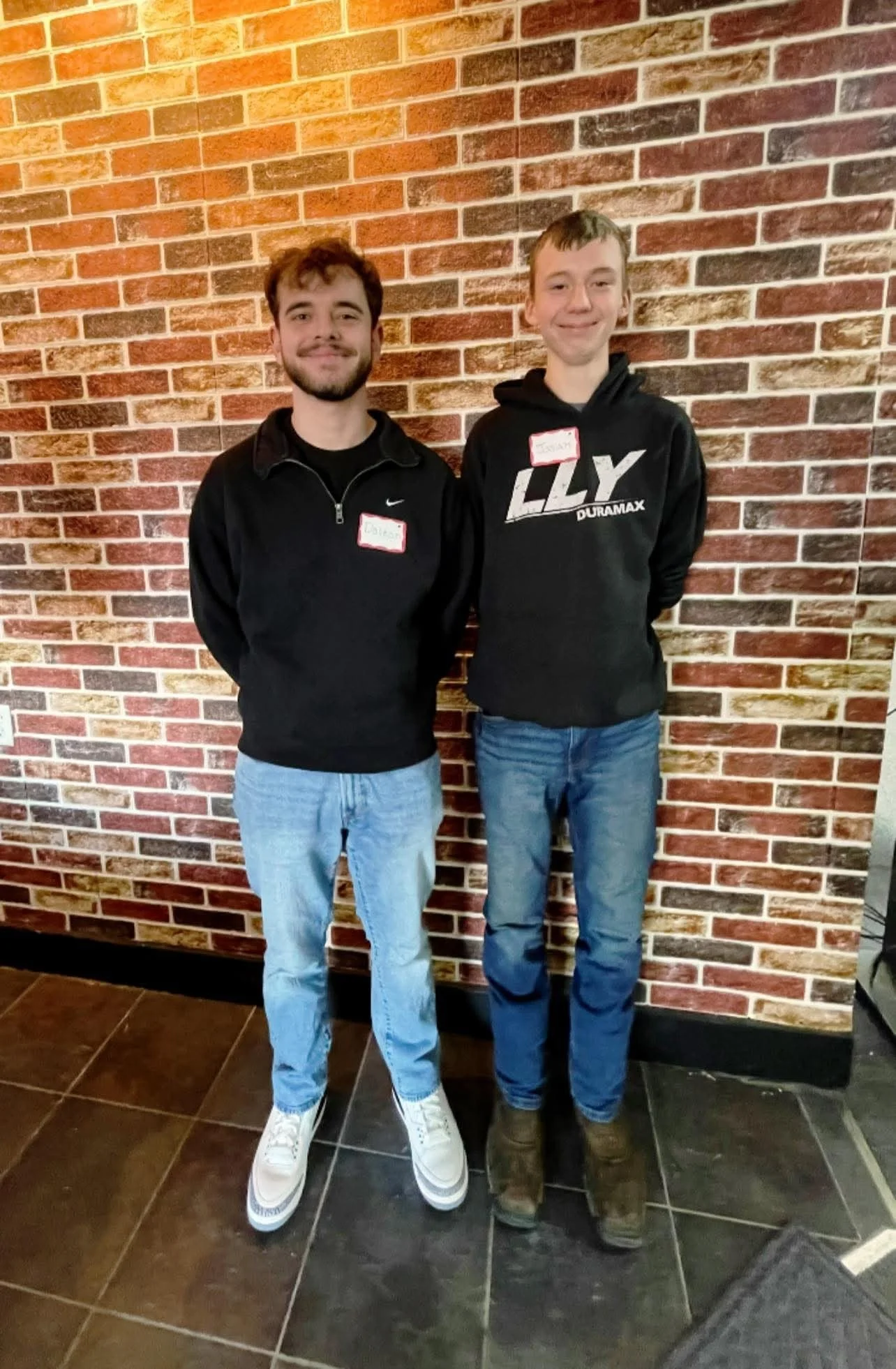 A mentor and a mentee standing next to each other against a brick wall. They are both smiling and wearing dark sweatshirts and jeans.