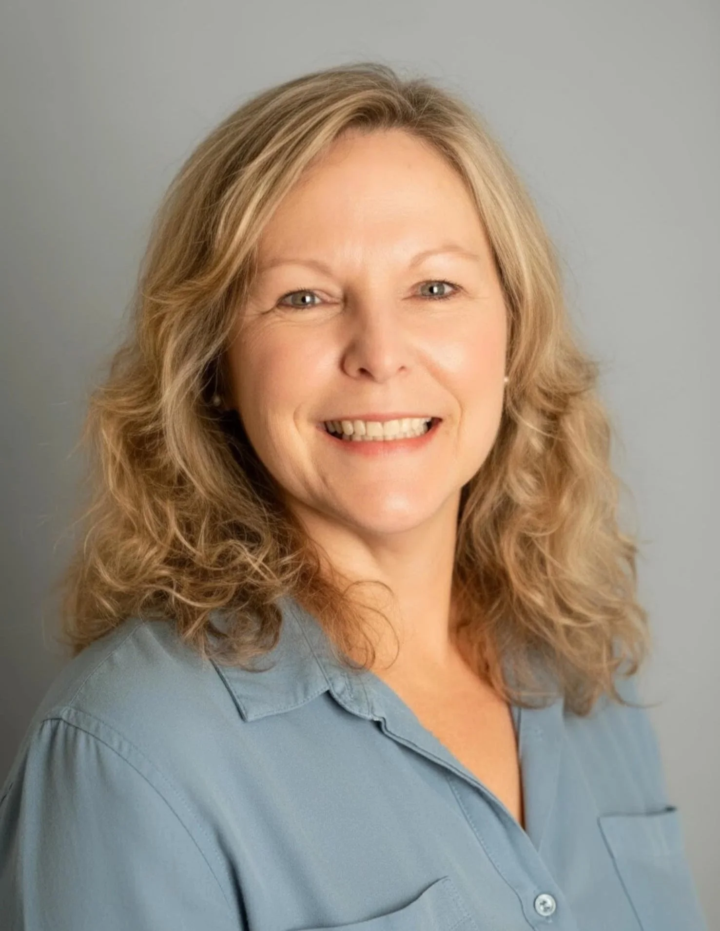 A headshot portrait of CCM Director, Victoria Caldwell–a woman with blonde, curly hair smiling, wearing a light blue button-up shirt, against a light gray background.