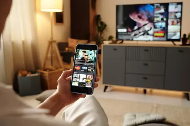 Person sitting on a couch holding a smartphone, watching a television in a cozy living room