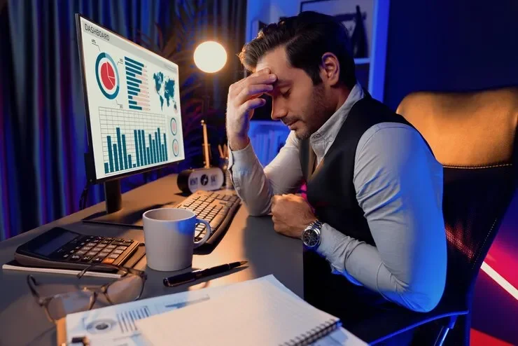 Man sitting at a desk with a stressed expression, hand on forehead, surrounded by a computer monitor with charts and graphs, a coffee mug, calculator, and documents.