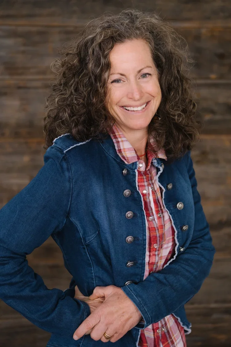 Lisa, a smiling woman with curly brown hair wearing a denim jacket over a plaid shirt, posing with one hand on her hip against a wooden wall background.