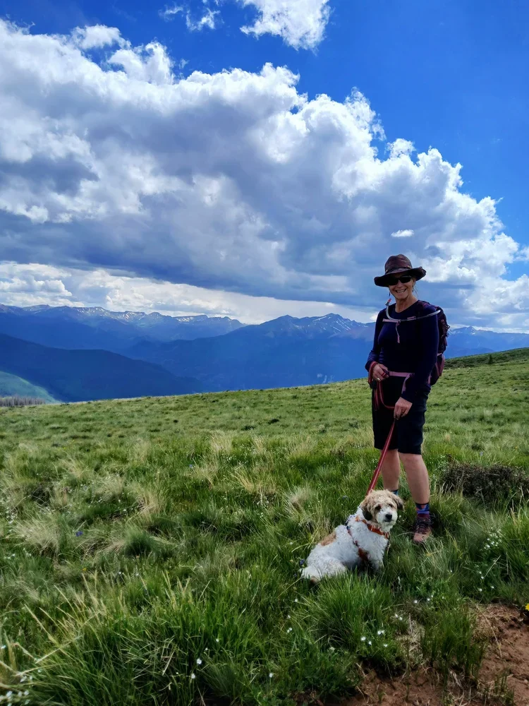 Lisa standing on a grassy hill with a small white and brown dog on a leash, mountains and a partly cloudy sky in the background.