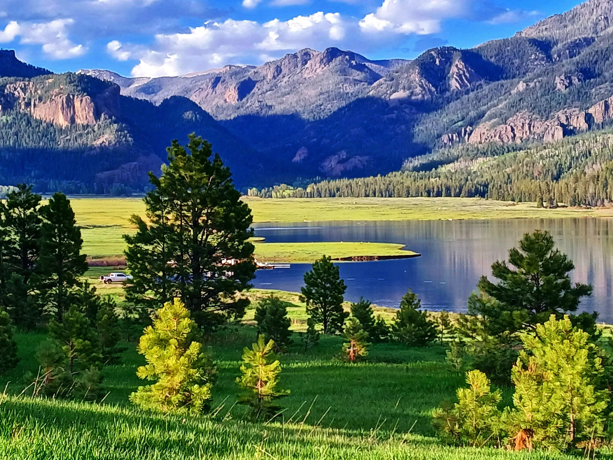 A scenic landscape of Williams Lake Reservoir, in Pagosa Springs, Colorado, showing a mountain range in the background with a cloudy sky, a body of water, and a lush green area with trees and grass in the foreground.
