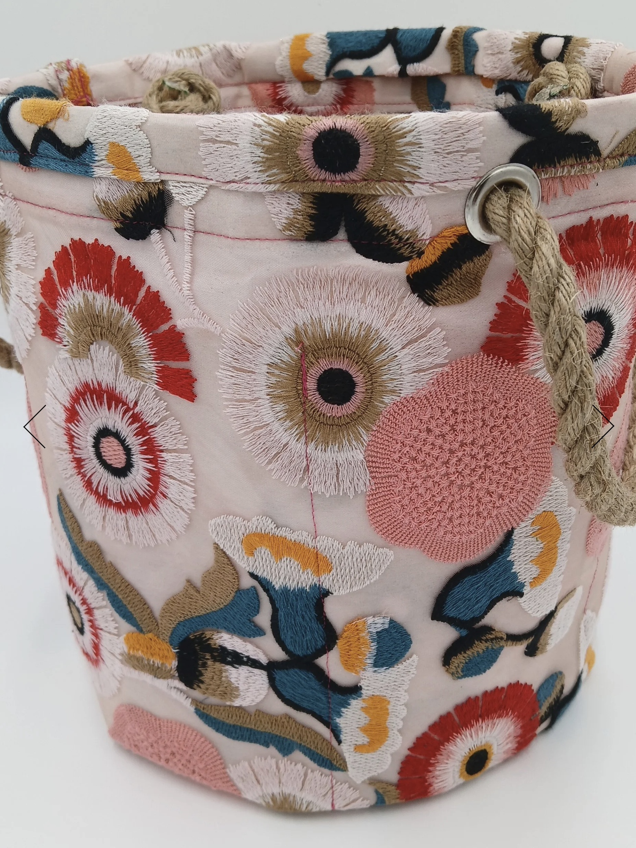 Close-up of a fabric bag with floral embroidery featuring colorful flowers with black centers, pink, red, white, blue, and yellow petals, and a rope drawstring threaded through a metal eyelet.