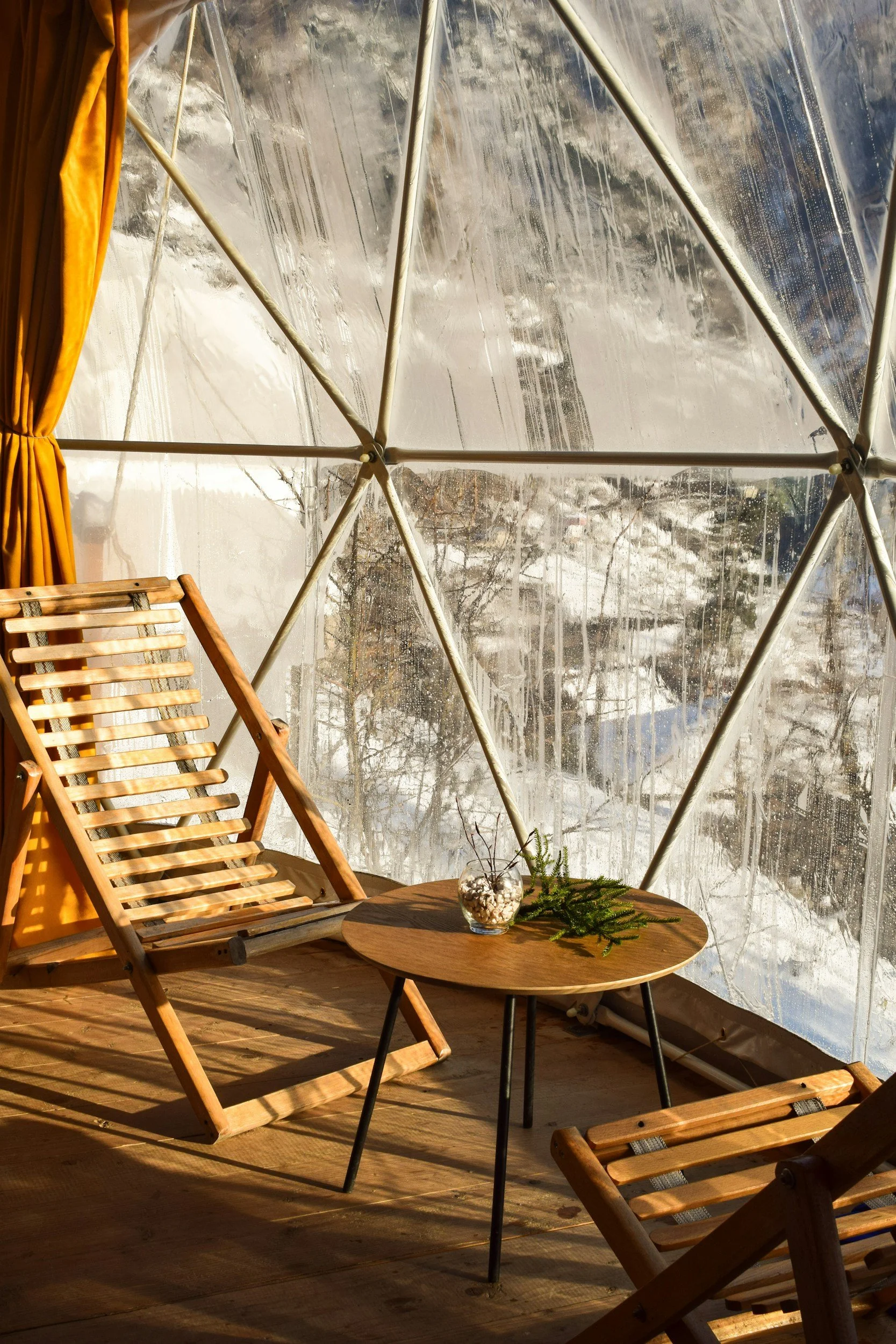 Indoor scene with wooden chairs, a small wooden table, and a glass bowl with decorative stones and pine branches, set inside a geodesic glass greenhouse with winter landscape outside.