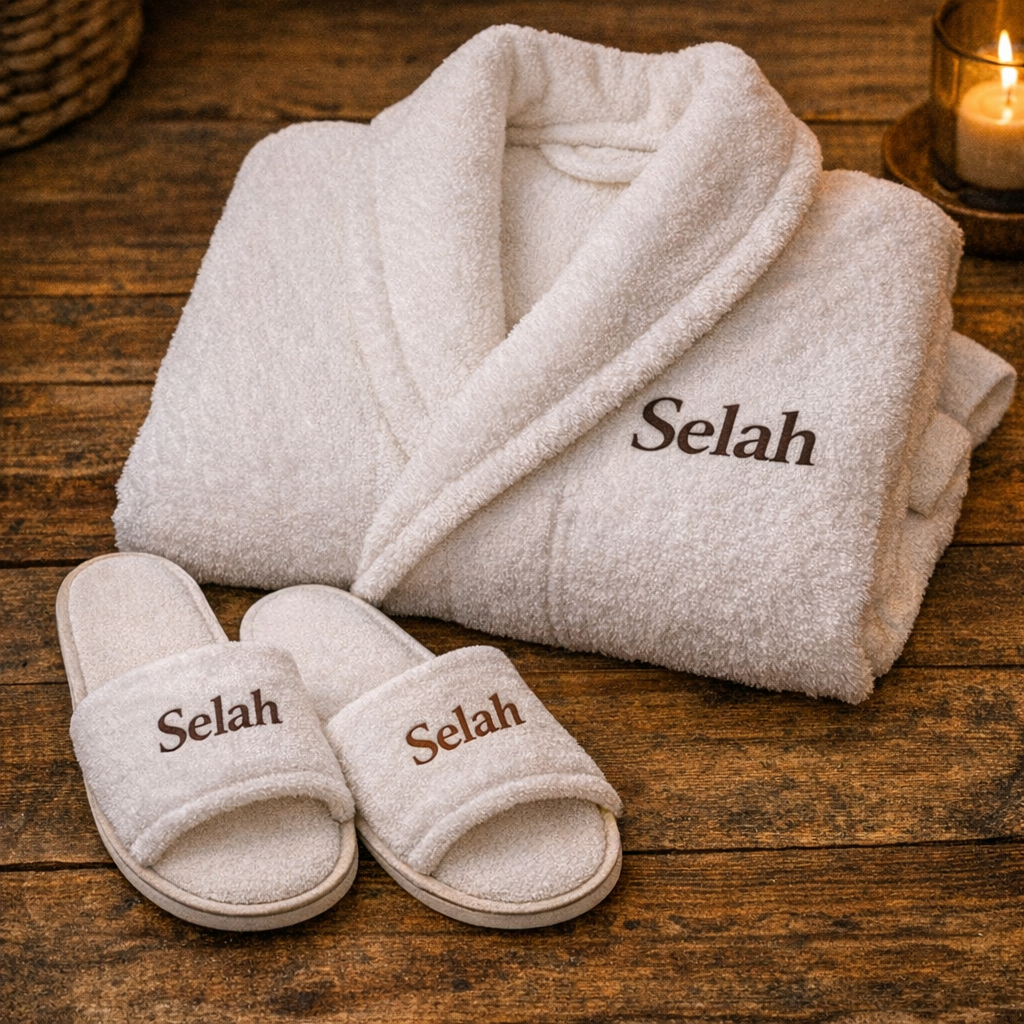 White towel with the name "Selah" embroidered on it, folded on a wooden surface next to a pair of matching slippers and a lit candle.