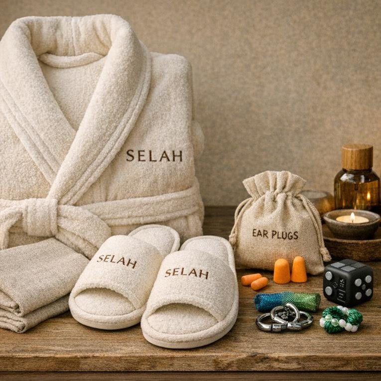 Spa and self-care items including a white bathrobe and slippers personalized with 'Selah', folded towels, a bag labeled 'ear plugs', orange and beige ear plugs, a roll of blue tape, a black die, some metal rings, and a bracelet made of green and white beads, all arranged on a wooden surface.