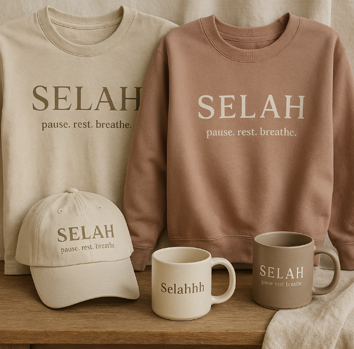 Display of beige and pink sweatshirts, a beige cap, and two coffee mugs with the logo 'SELAH' and the phrase 'pause. rest. breathe.' on a wooden surface.