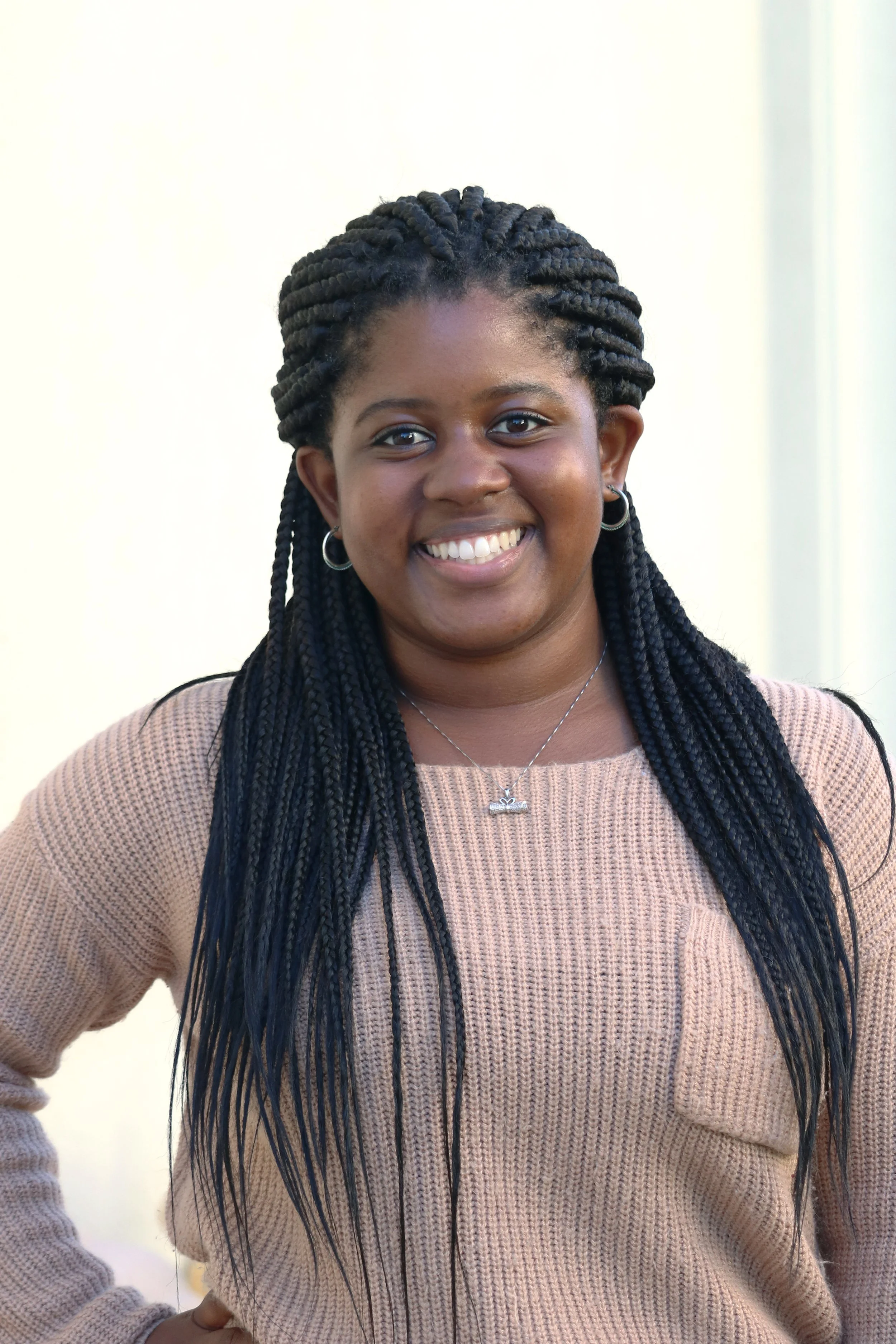 Portrait of a young woman with dark braided hair wearing hoop earrings and a beige knit sweater, smiling at the camera.