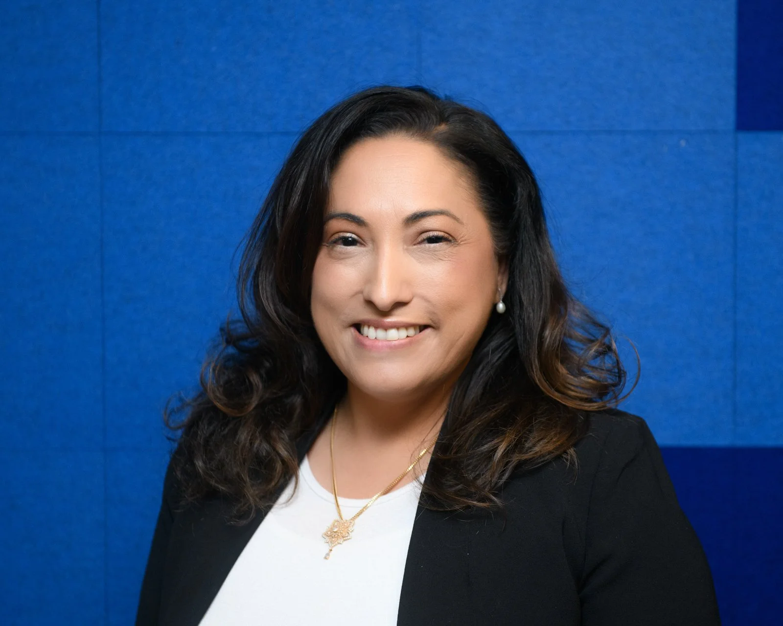 A woman with black, wavy hair is smiling at the camera, wearing a black blazer, white top, pearl earrings, and a gold necklace with a pendant, standing against a blue background.