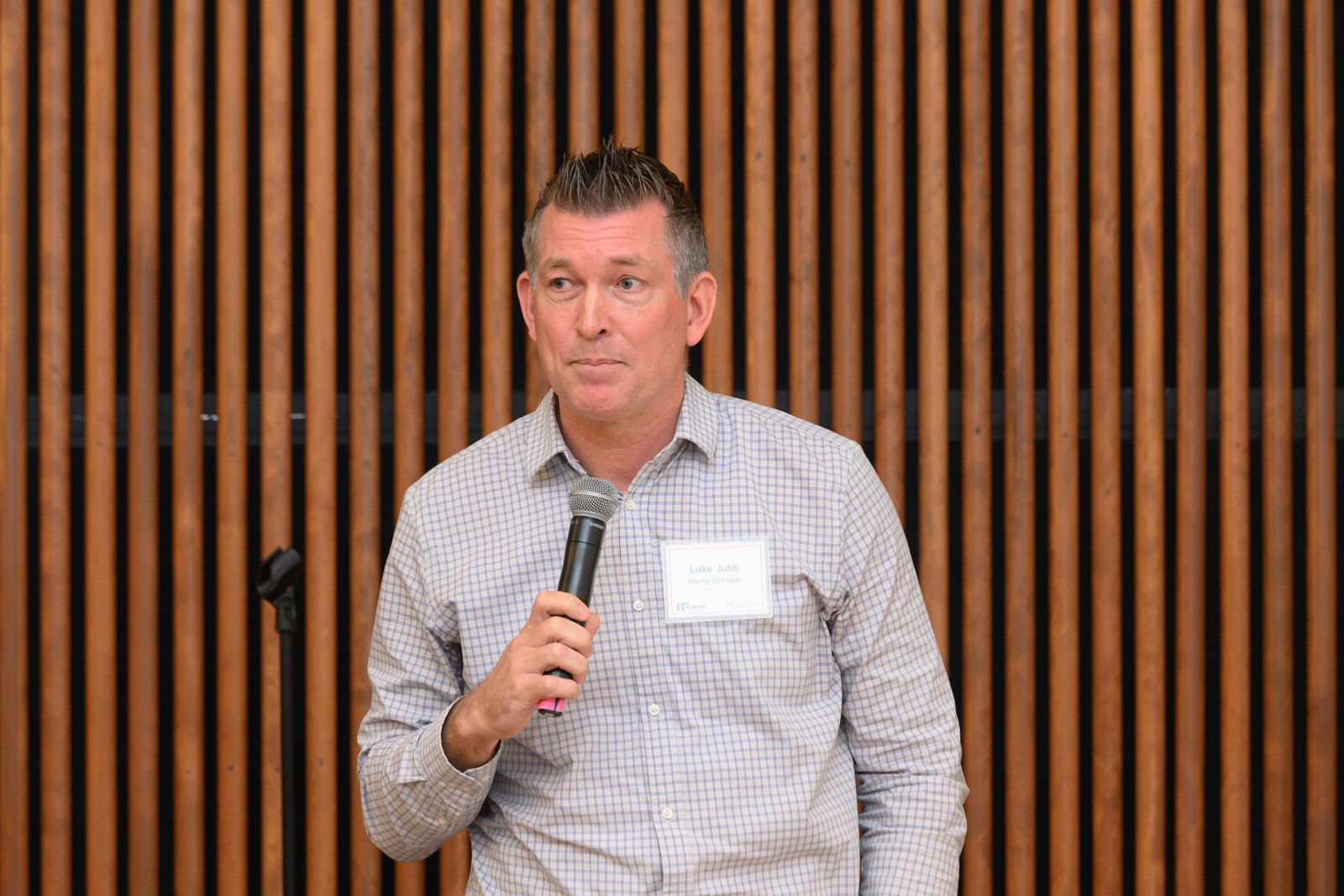 A man with short dark hair and a checkered shirt is holding a microphone and speaking at an event with a wooden slat wall in the background.