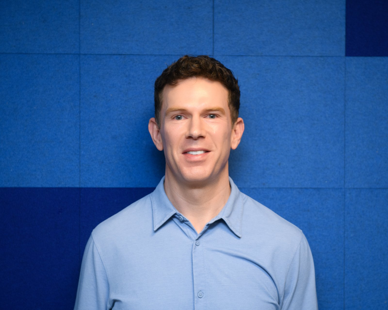 Portrait of a man with short brown hair wearing a light blue button-up shirt, standing in front of a blue padded wall.