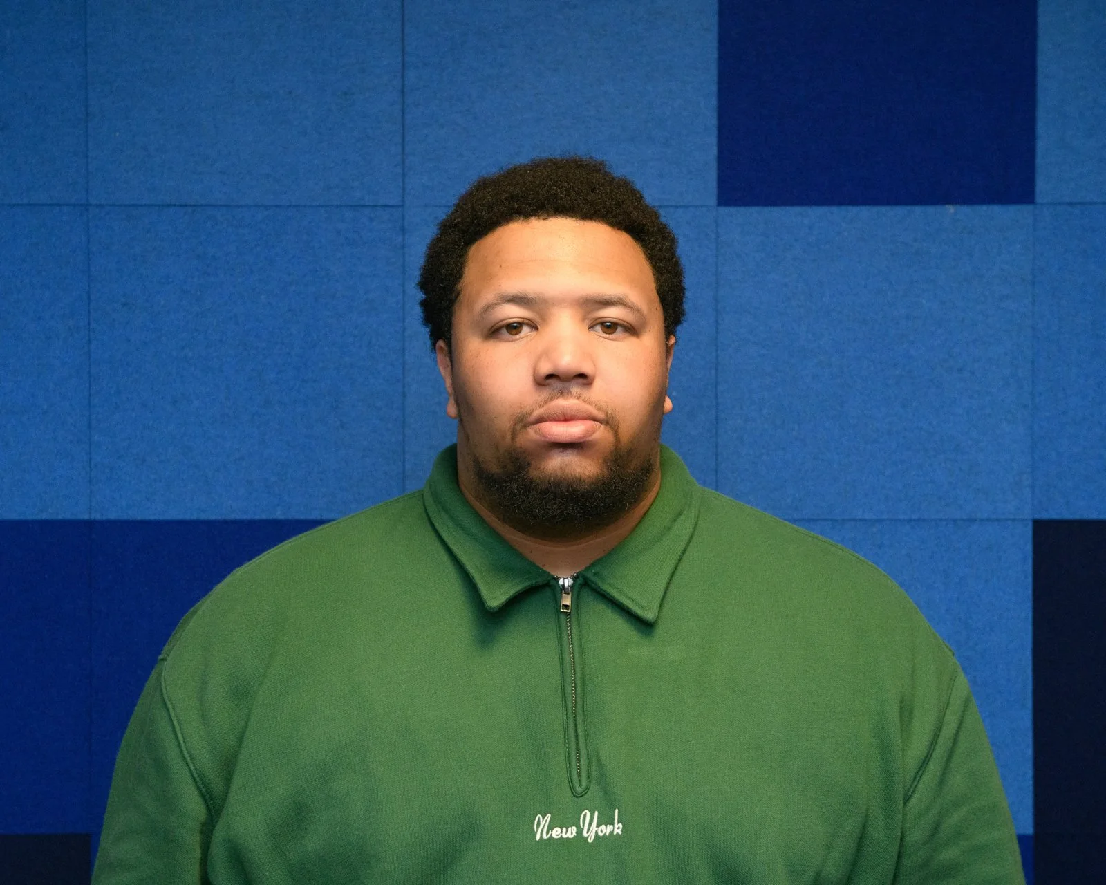 A man with curly hair and a beard wearing a green zip-up jacket with 'New York' embroidered on it, standing against a blue tiled wall.