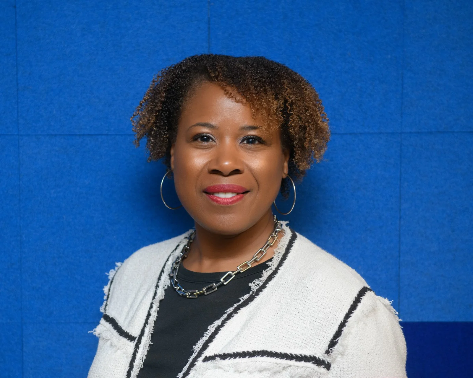 Portrait of a woman with curly hair, wearing hoop earrings, a chain necklace, and a black top with a white and black jacket, standing against a blue background.