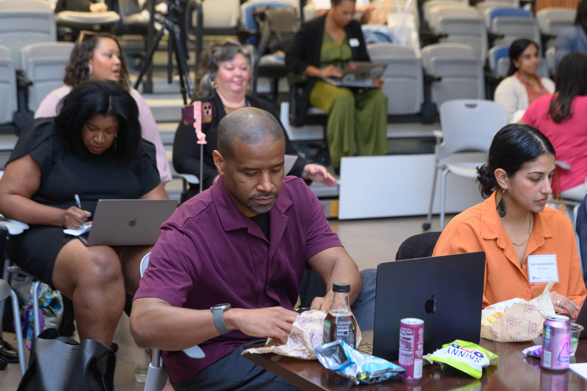 People sitting at tables with snacks, drinks, and laptops in a conference or workshop setting, some engaged with their devices, others looking around or talking.