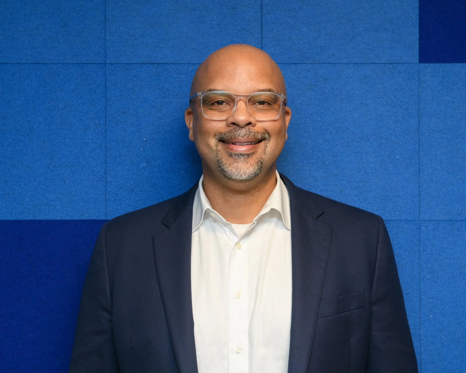 A man with glasses, a beard, and a mustache is smiling and dressed in a suit and white shirt, standing against a blue wall.