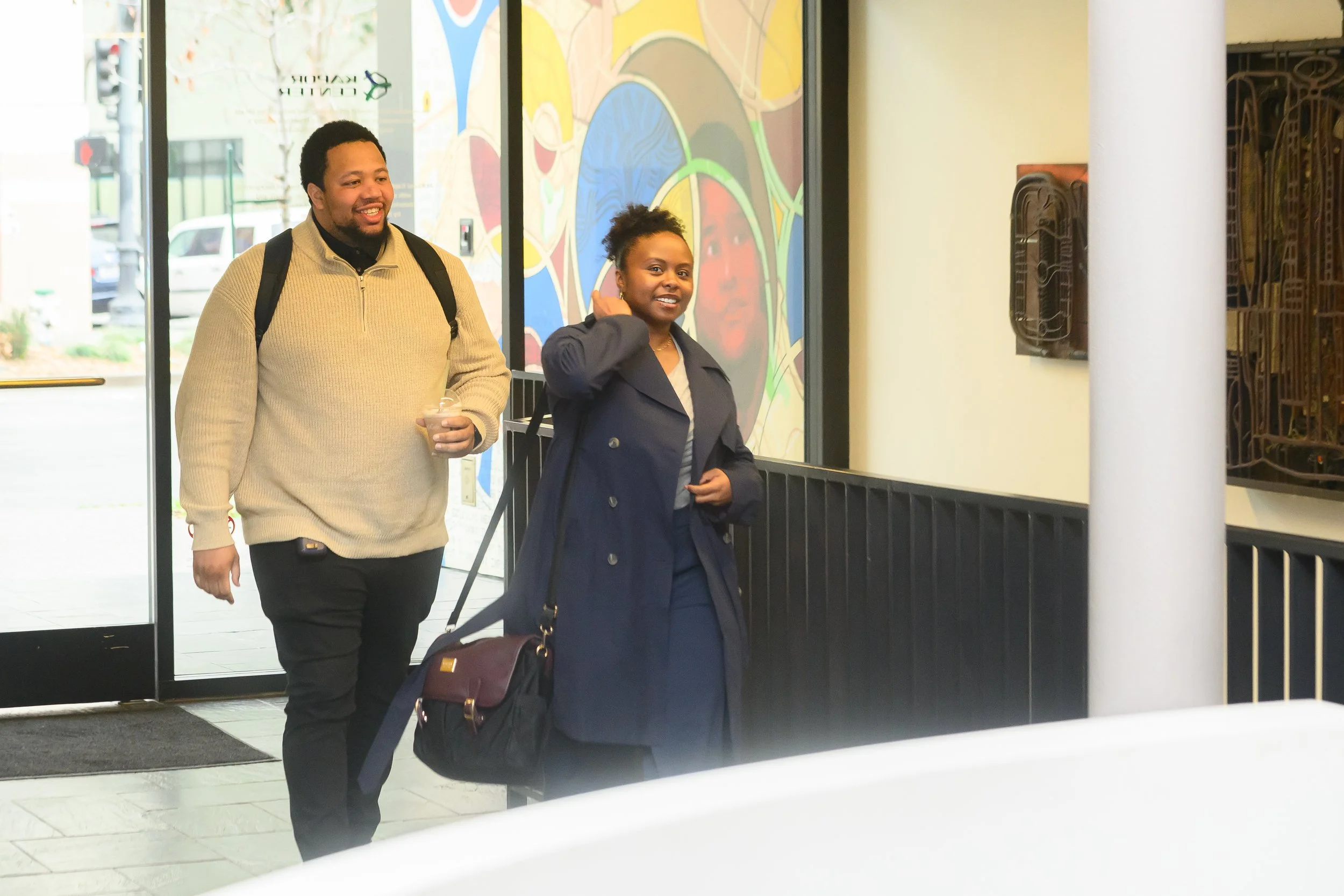 A man and a woman walking out of a building, smiling, with a colorful mural on the wall behind them. The man is carrying a drink and wearing a beige sweater, while the woman is adjusting her earring and wearing a dark blue coat.