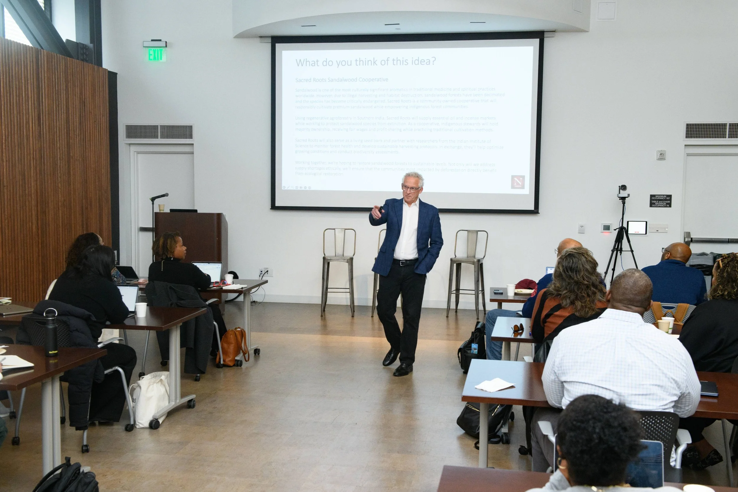 A man in a blue suit giving a presentation to an audience in a conference room. The presentation slide on the screen reads 'What do you think of this idea?' and discusses sacred roots sandalwood cooperative. Several people are seated at tables, some 