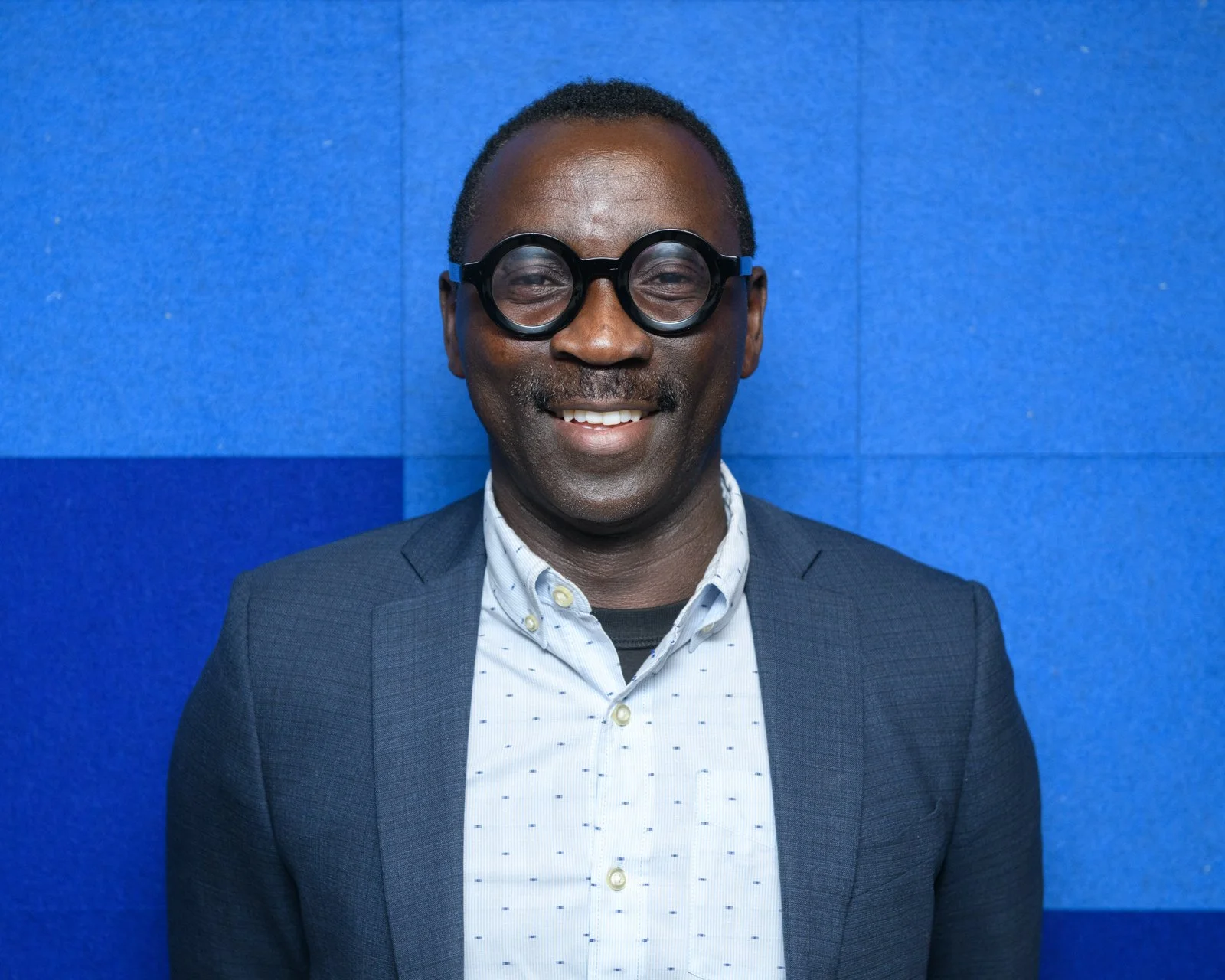 A smiling man wearing round black glasses, a dark blazer, and a light-colored, patterned button-down shirt standing against a blue wall.