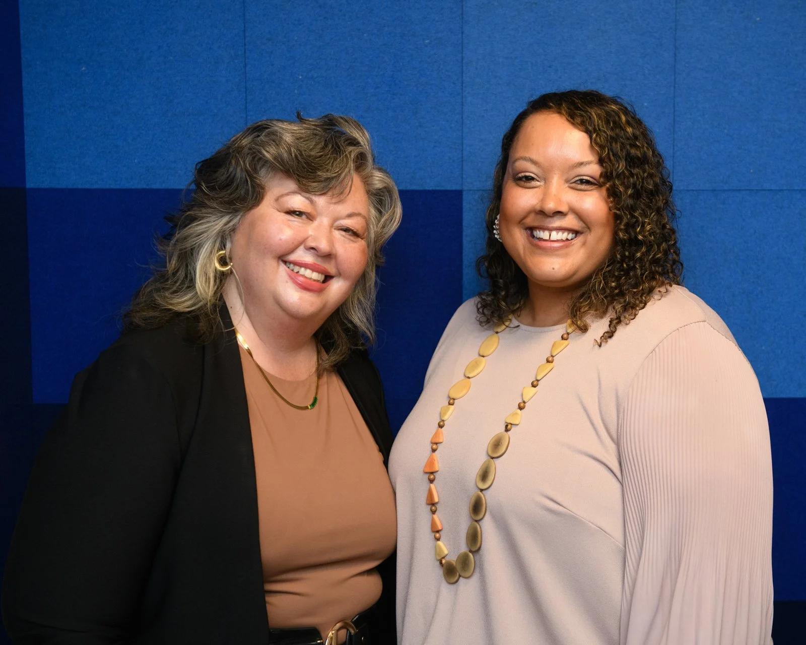 Two women standing side by side, smiling, in front of a blue wall.