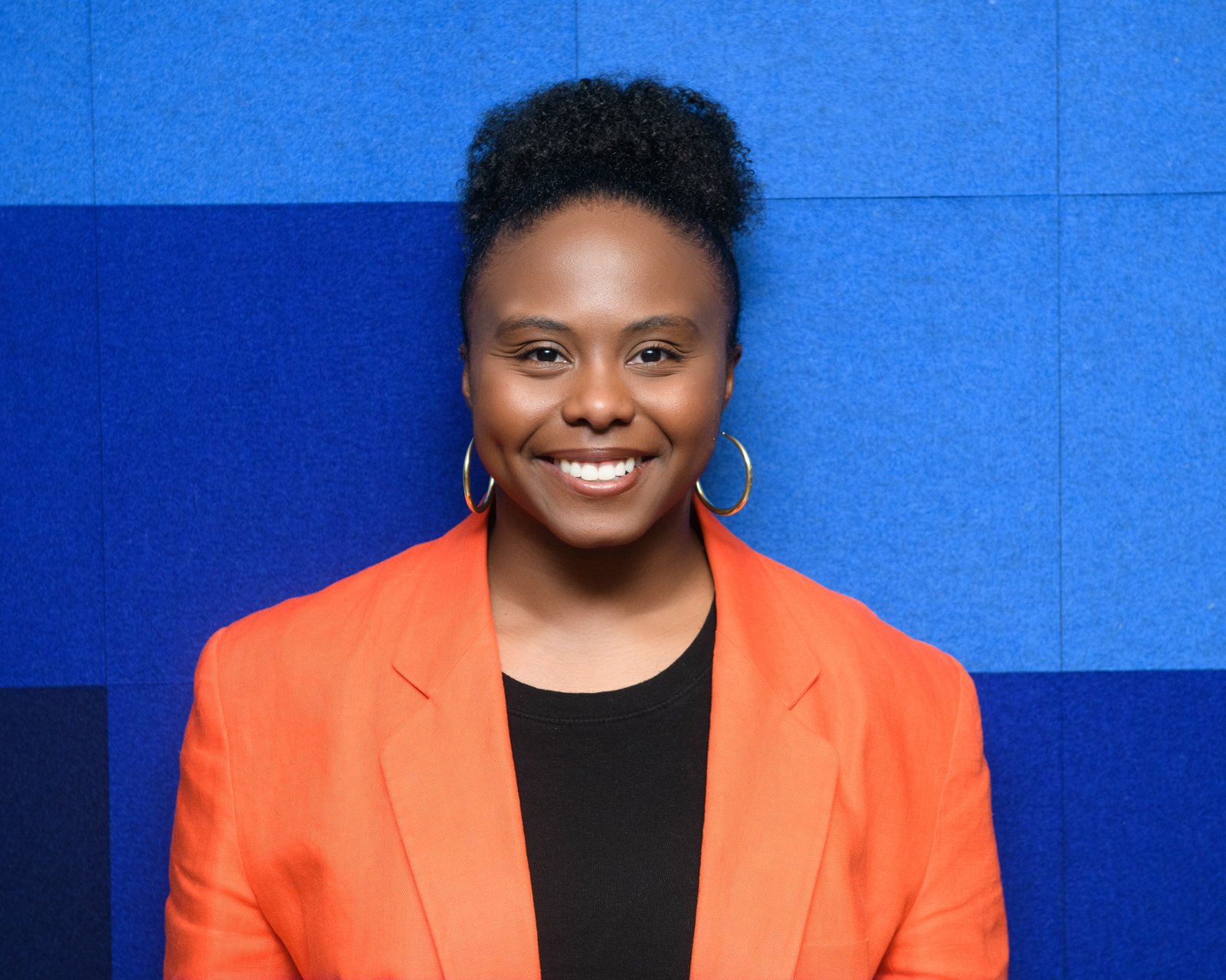 A woman smiling, wearing an orange blazer, black shirt, and hoop earrings, standing against a blue textured wall background.
