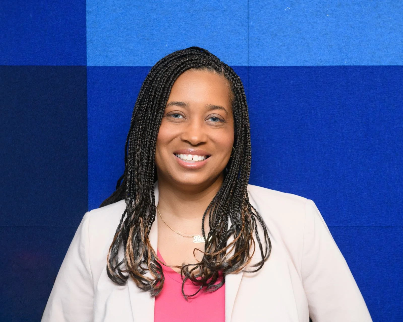 Portrait of a woman with black braided hair, smiling, wearing a white blazer over a pink top, standing against a blue geometric background.