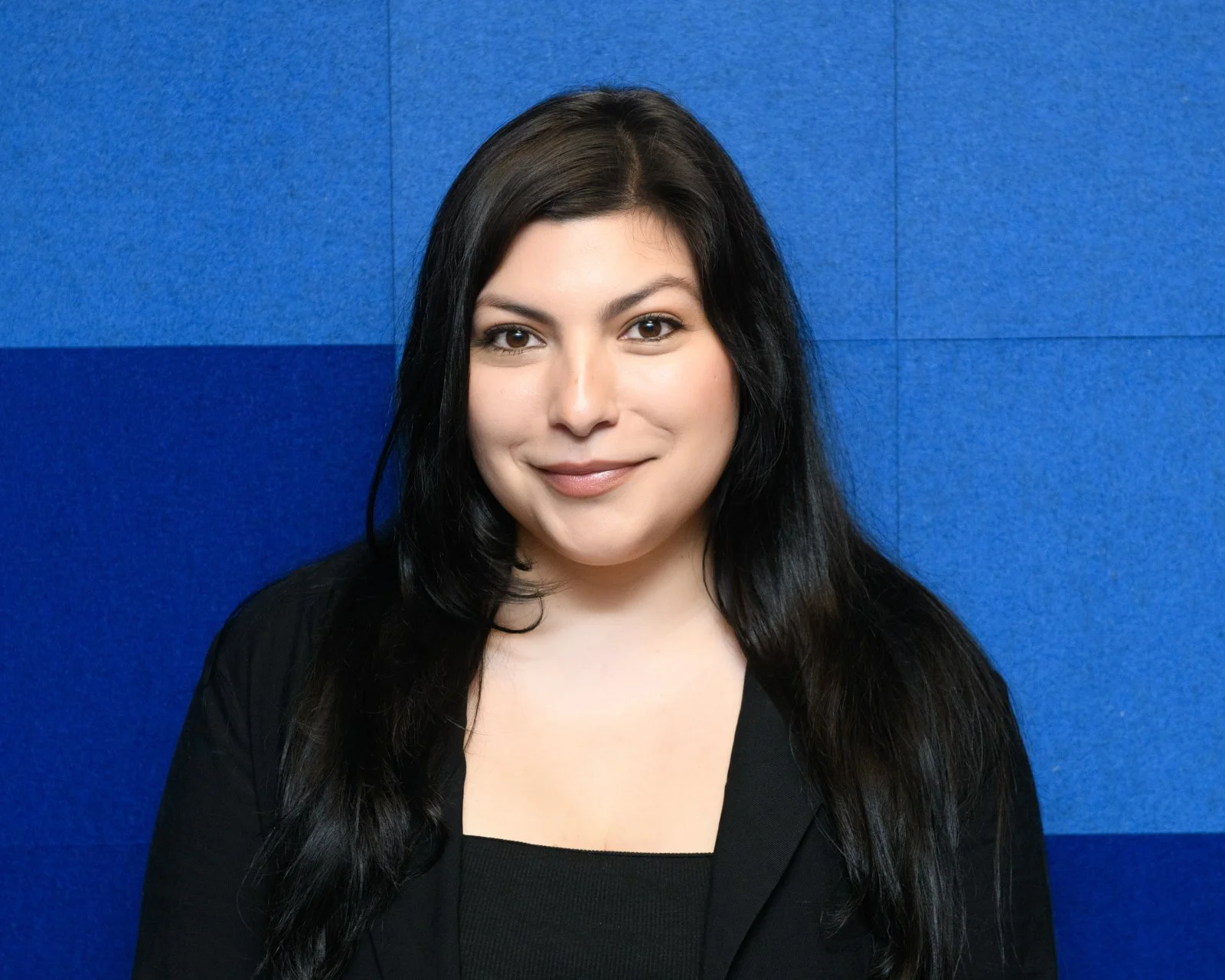Headshot of a woman with long dark hair, wearing a black top, standing against a blue textured wall.