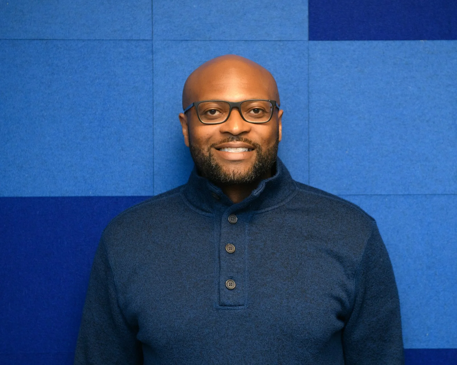A smiling man with glasses, a beard, wearing a dark blue button-up sweater, standing against a blue wall background.