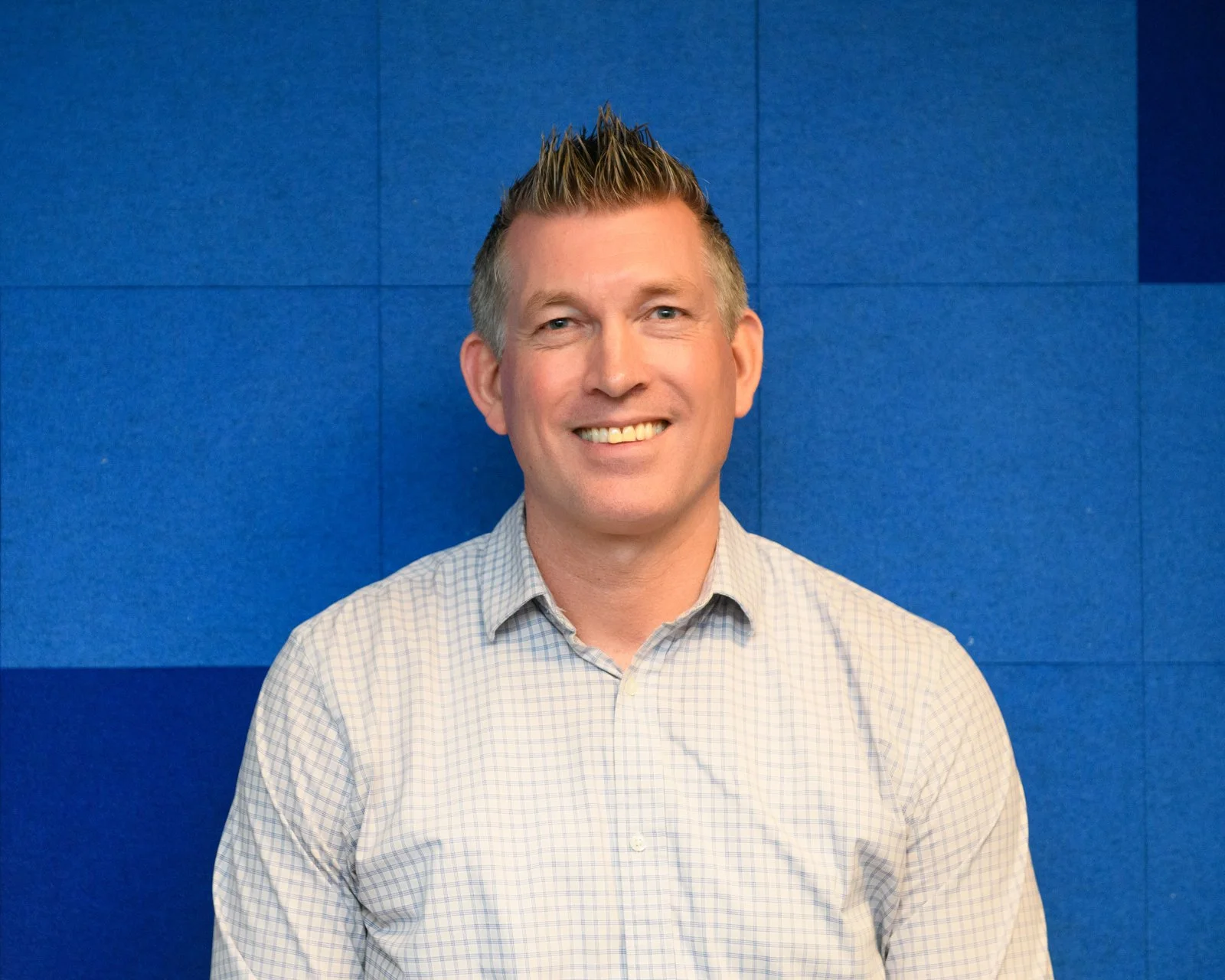 A smiling man with short, spiked hair wearing a light checkered shirt, standing in front of a blue wall with textured panels.