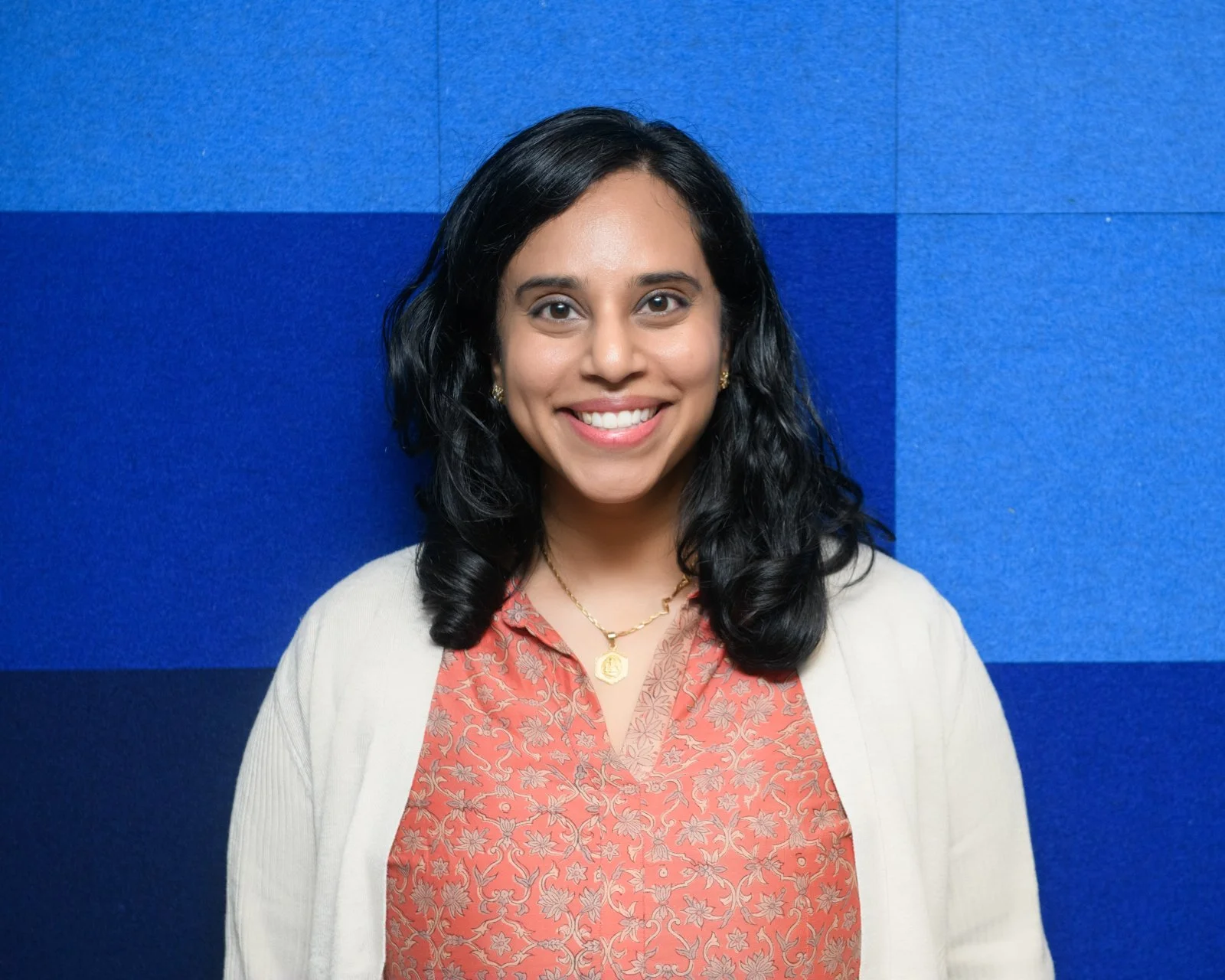 Portrait of a woman with black hair, smiling, wearing a coral-patterned blouse, gold jewelry, and a white cardigan, standing in front of a blue wall.