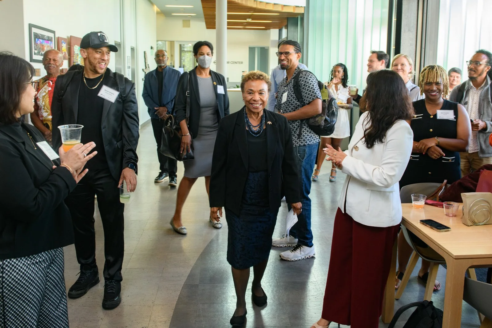 A diverse group of people in professional attire gathering in a brightly lit office space, engaging in conversation and smiling. Some are holding drinks, and one woman in the center walks through the crowd smiling.