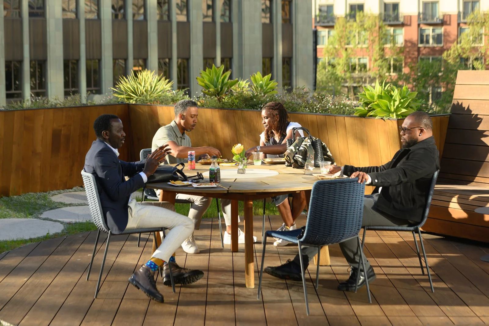 Four people having a meal outdoors at a round table on a rooftop patio with greenery and city buildings in the background.