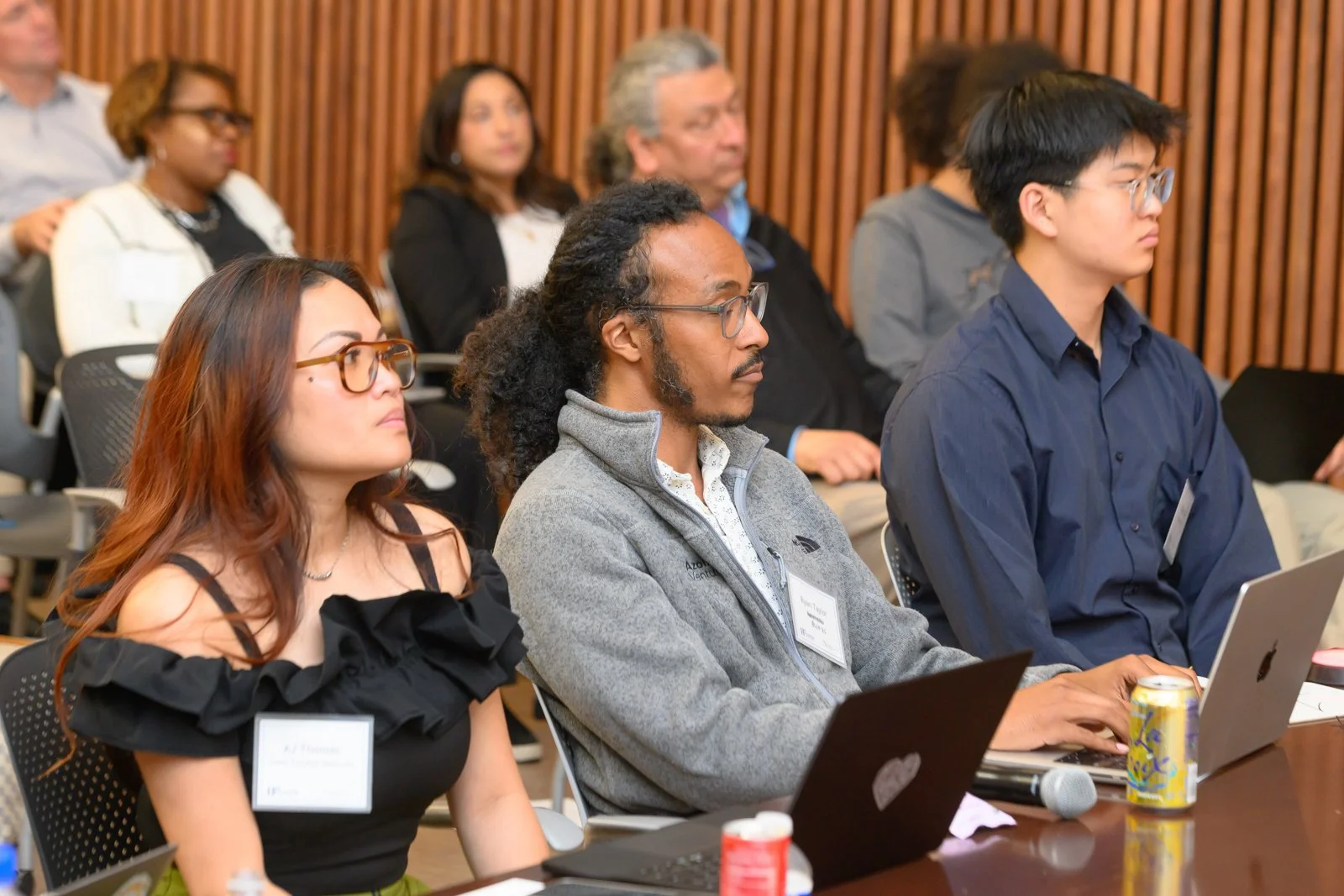 A diverse group of people attending a conference, sitting with laptops and notebooks, paying attention to a speaker in a room with wooden panel walls.