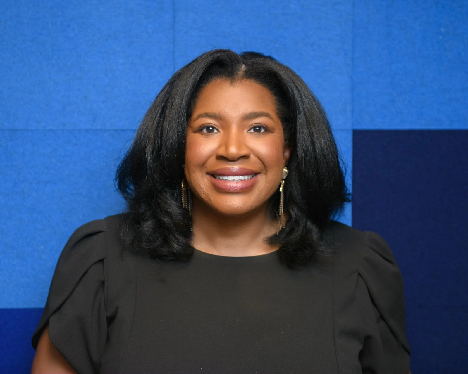 A smiling African American woman with shoulder-length black hair, wearing a black top with puffed sleeves and pearl earrings, standing in front of a blue textured wall.