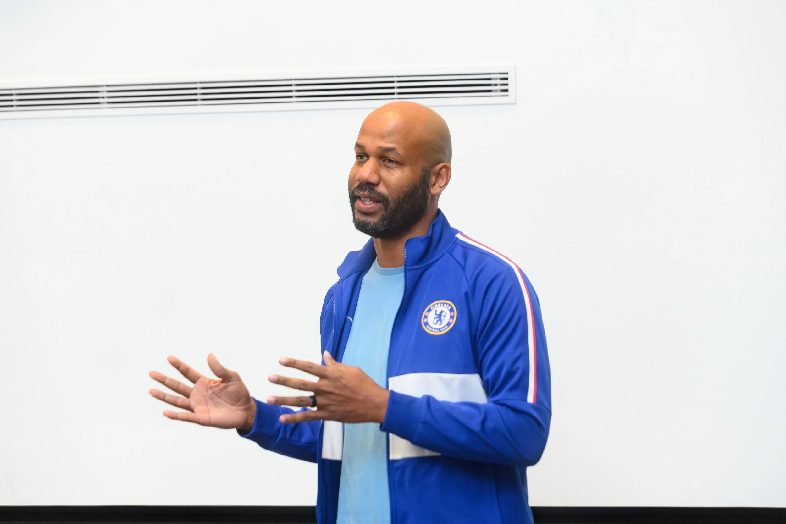 A bald man with a beard wearing a blue Chelsea football jacket speaking in front of a plain white wall.