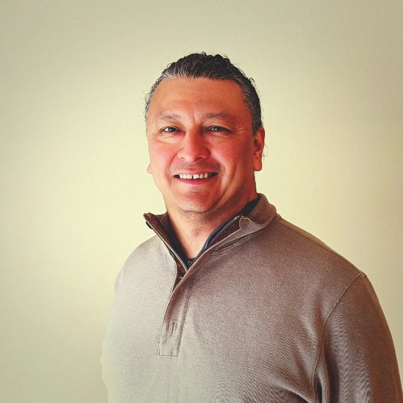 A smiling man with short, wavy dark hair, wearing a brown zip-up sweater, standing against a plain light-colored background.