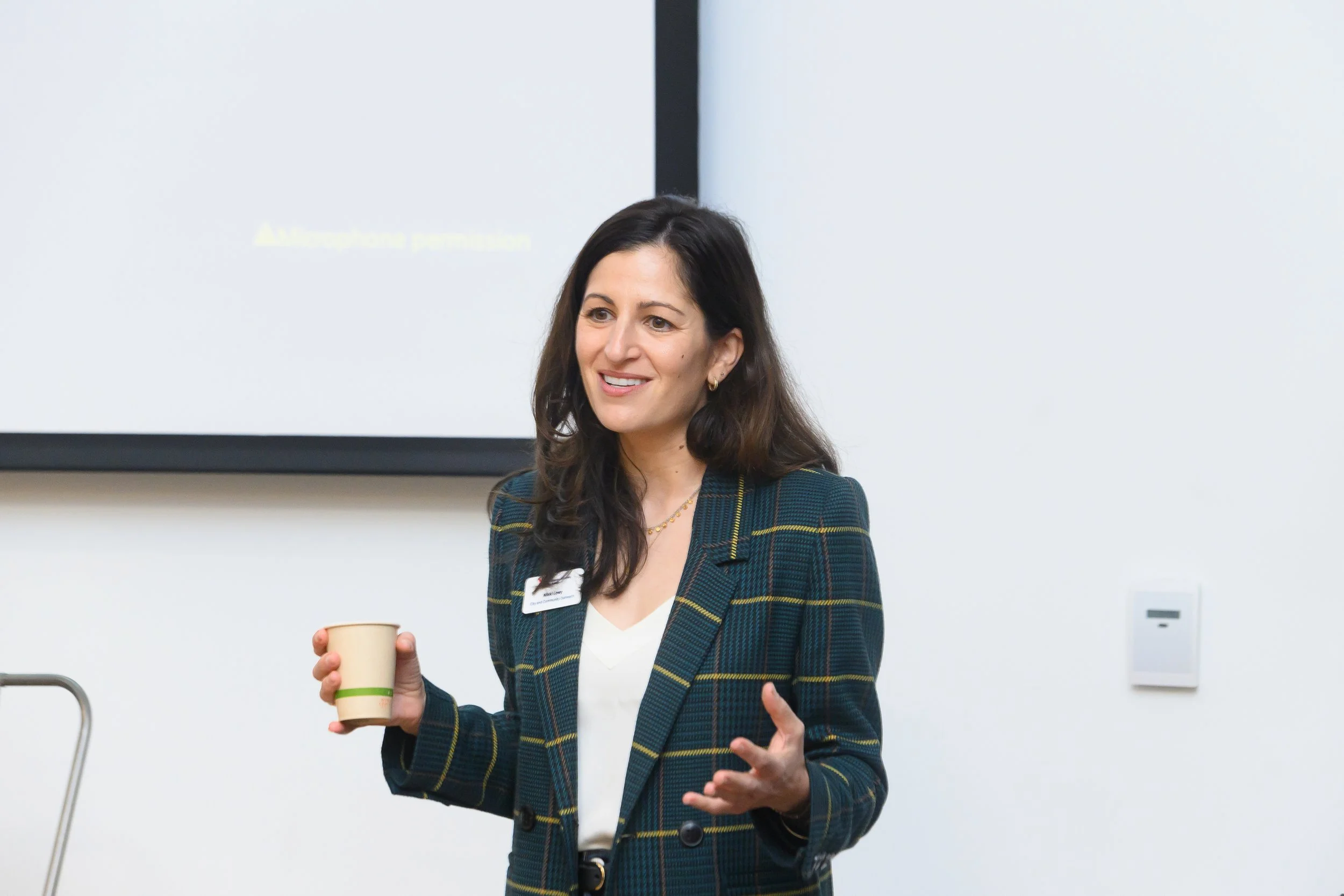 A woman with long dark hair, wearing a dark green checked blazer and a white top, is holding a paper coffee cup in her right hand while speaking in a classroom or conference room setting.